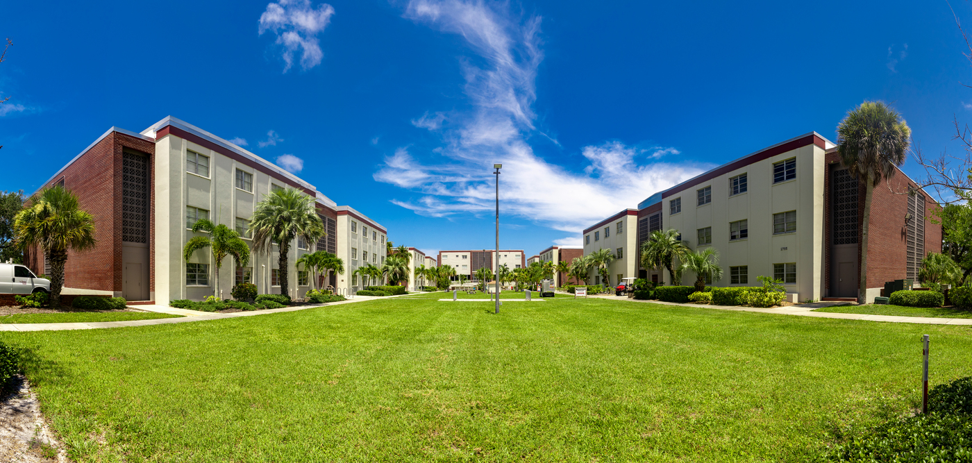 A large open quad with green grass on a bright sunny day. Three story buildings are seen around the perimiter of the quad.