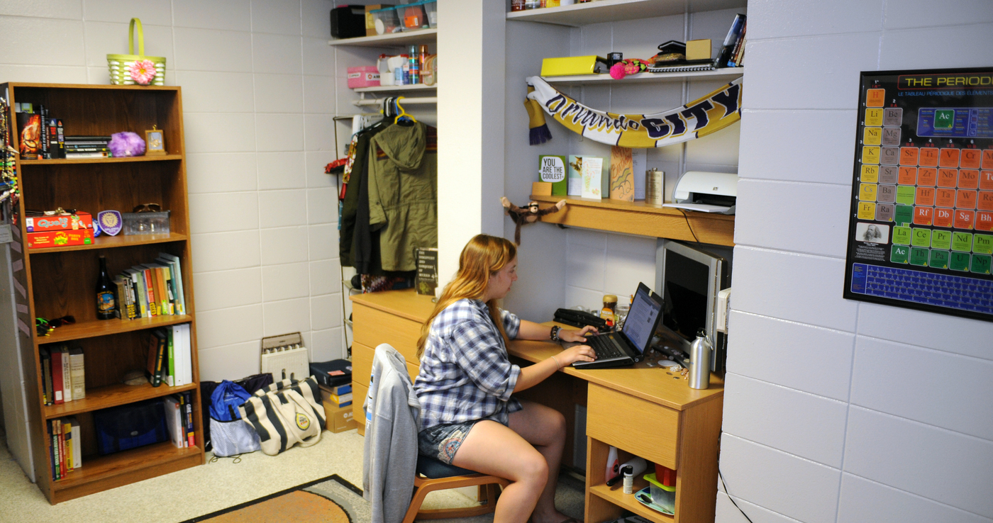 A student sits at a desk in their dorm room. Shelves full of books and supplies are above the desk and a dresser, bookshelf and closet space is visible in the background.