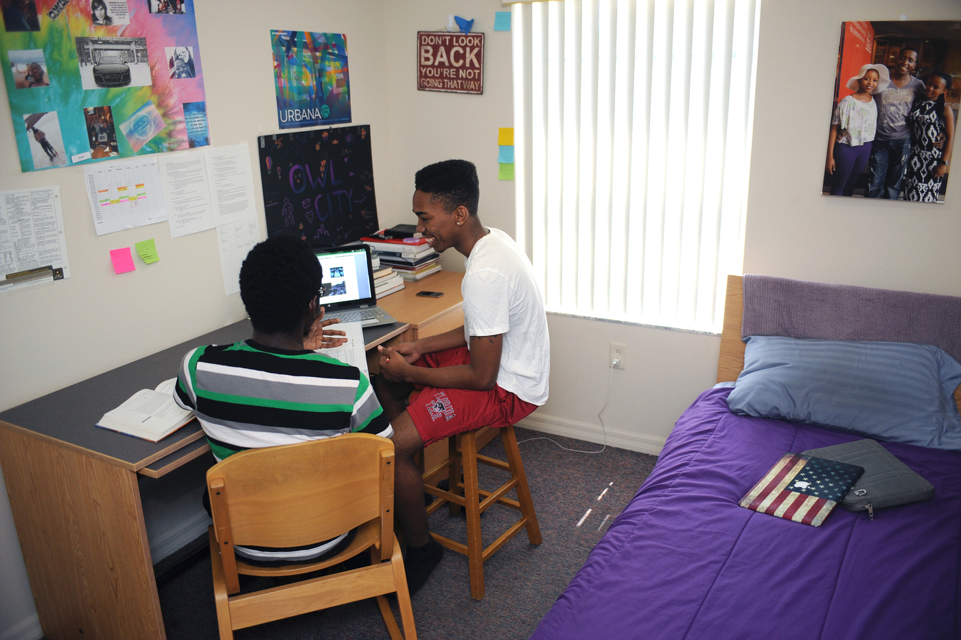 Two students having a conversation around a laptop on a desk in a dorm room. Posters hang on the walls and bed with bright colored bed spread is visible to one side of the room.