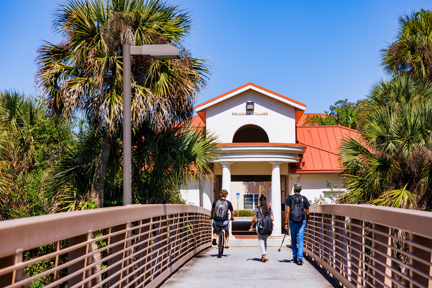 Columbi Village pedestrian bridge surrounded by dense foliage. Columns on either side of an entryway to the community common building is visible at the end of the bridge