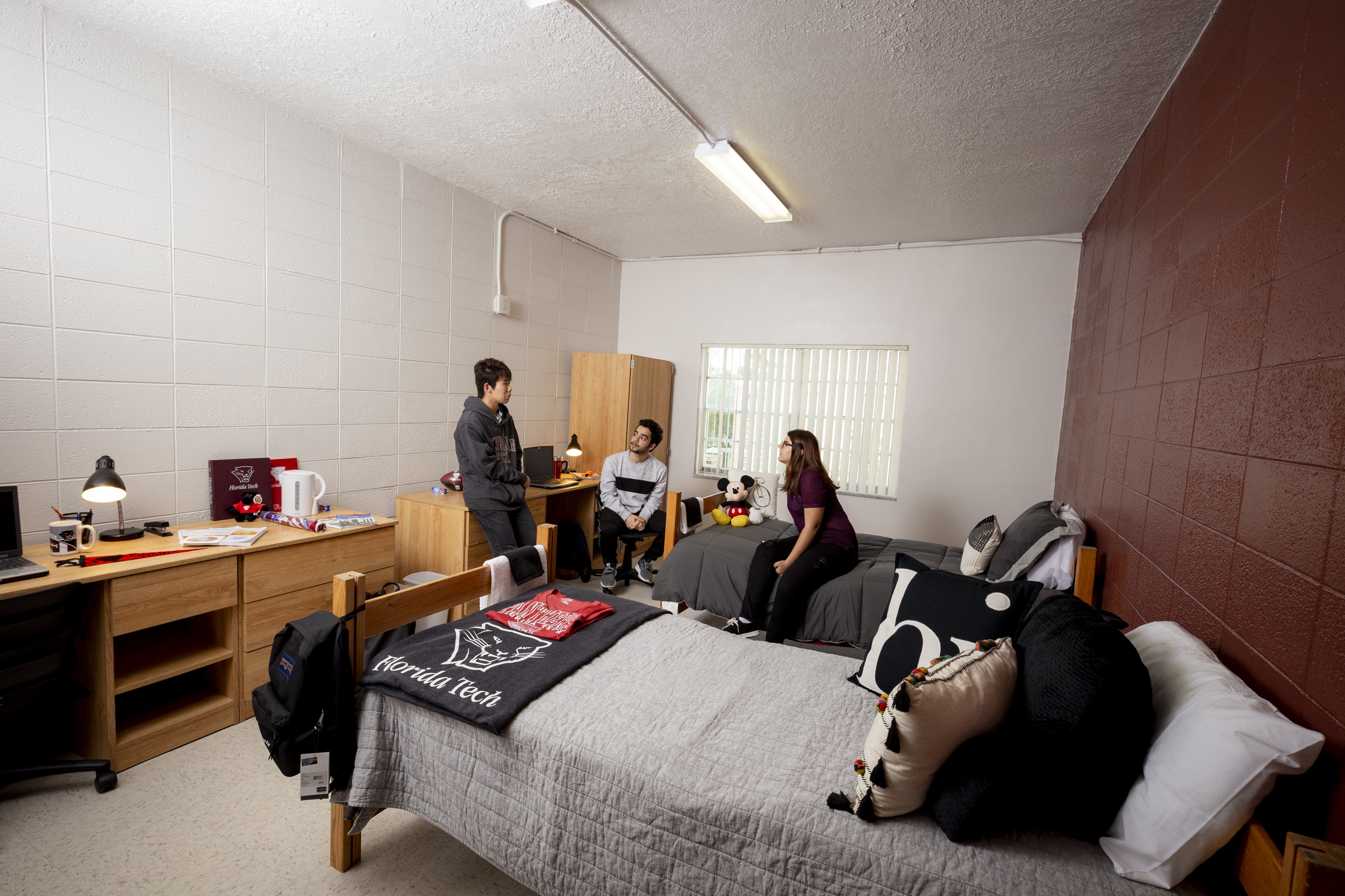 3 Students laughing having a conversation in a dorm room. Two beds, desks, dressers and wardrobes are visible in the room