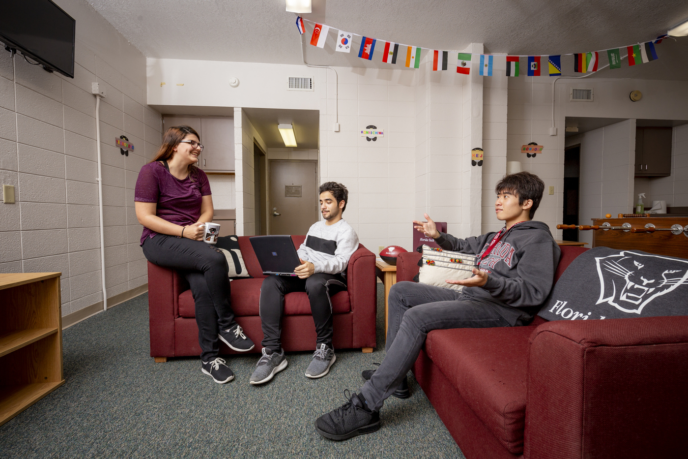 Three students hanging out in a residence hall common area. A TV, foosball table, and couches are visible