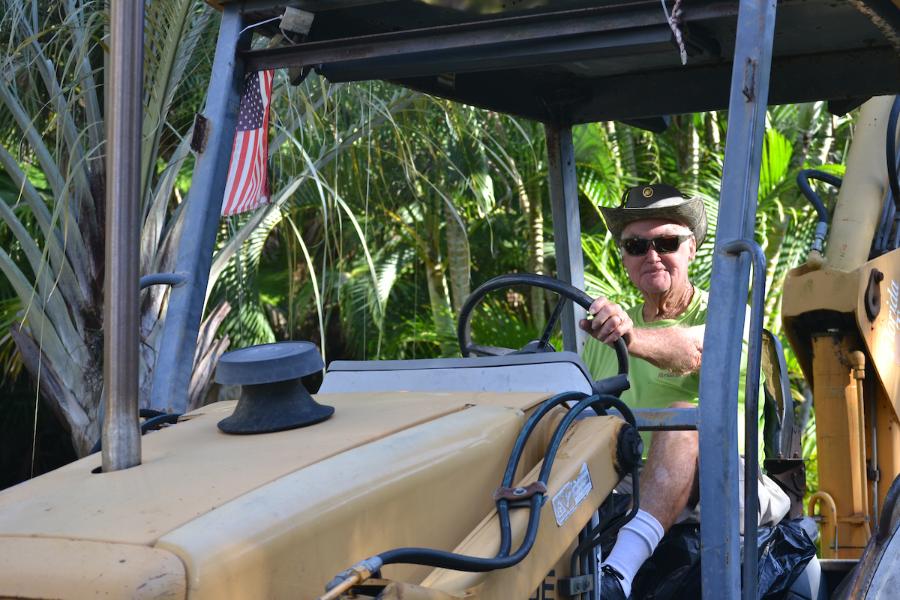 A friendly-looking, mature groundskeeper, Hank Hughes, in a neon green t-shirt, cowboy hat and sunglasses, driving a large tractor.